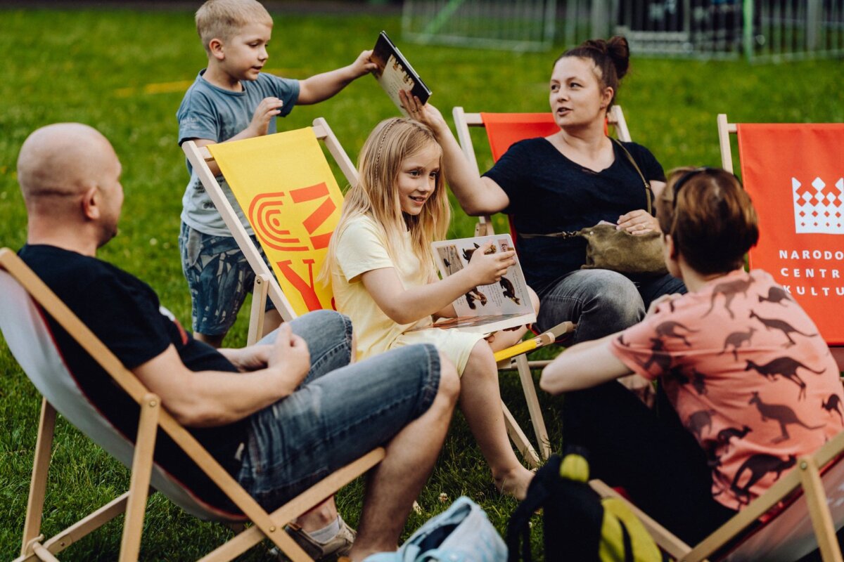 A group of people, including adults and children, are sitting in deck chairs outdoors on a grassy area. One child holds an open picture book, while another shows it to an adult. There are bright deckchairs and a relaxed, casual atmosphere - perfect for a Photo Event.  