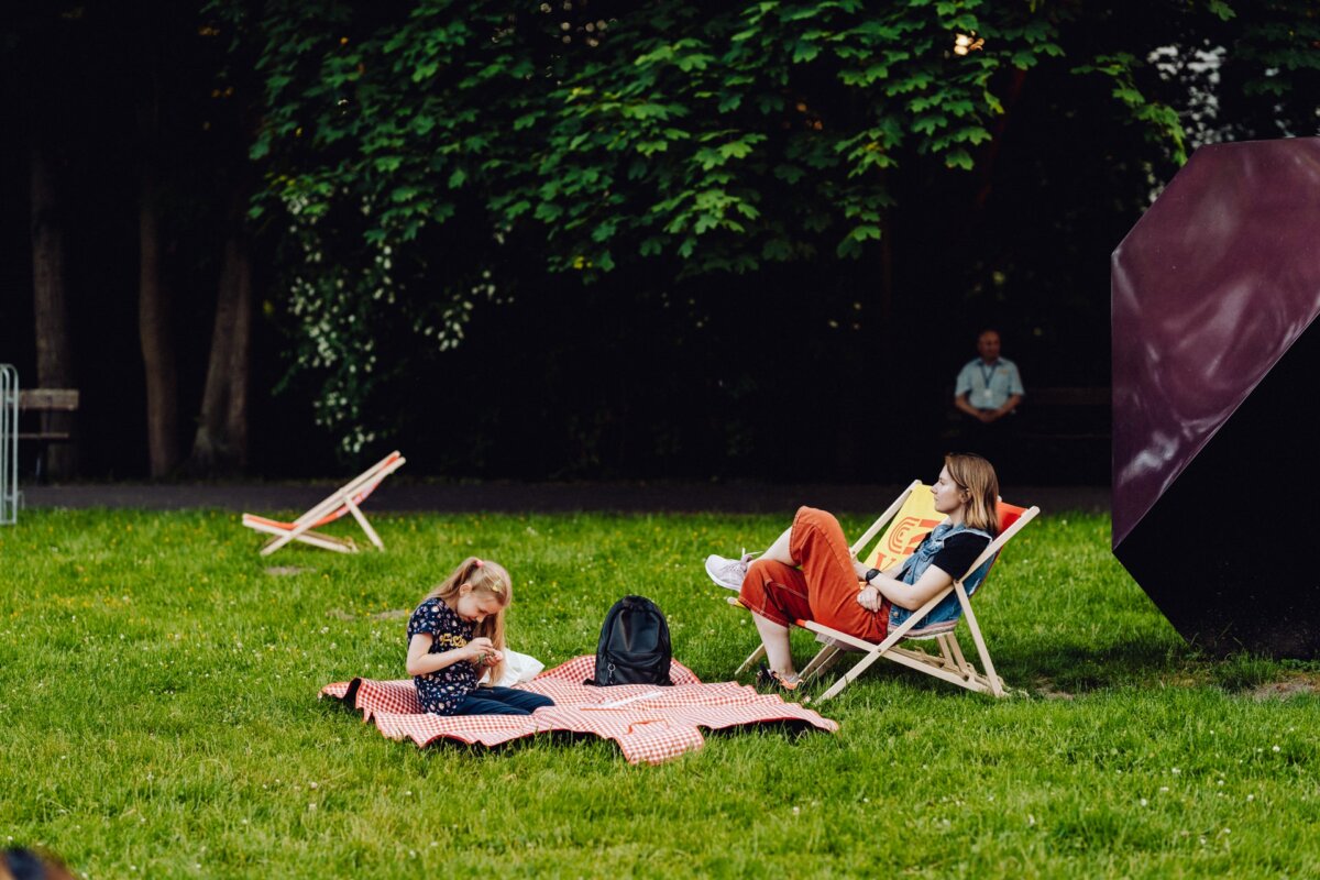 Two people are relaxing in the park: a woman sits on a deck chair and reads a book, and a child sits on a red and white checkered picnic blanket and plays. Next to them, a backpack is placed on the grass. In the background, among the lush greenery of the trees, additional deckchairs are visible, beautifully captured by our photographer from Warsaw.  