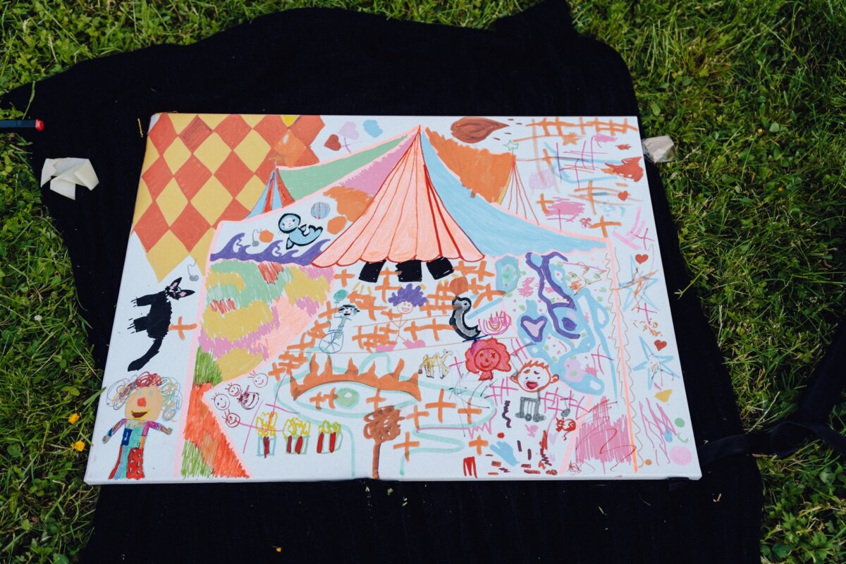 A colorful abstract painting on white canvas lies on a black canvas spread on grass. The painting features a variety of fanciful elements, including a circus tent, checkerboard patterns and doodles depicting people, animals and shapes in vibrant colors. Perfect for a Warsaw photographer to capture at any event.  