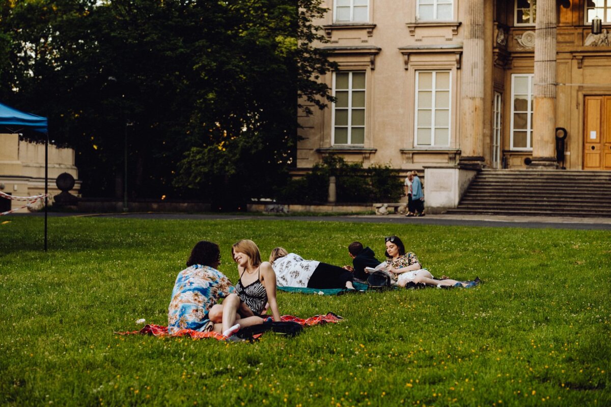 People are sitting in groups on blankets on the lawn near a large building that resembles a historical or academic institution. The atmosphere is relaxed, with some people talking and others relaxing. The scene, beautifully captured by event photographer Warsaw, takes place in the early evening or late afternoon.  