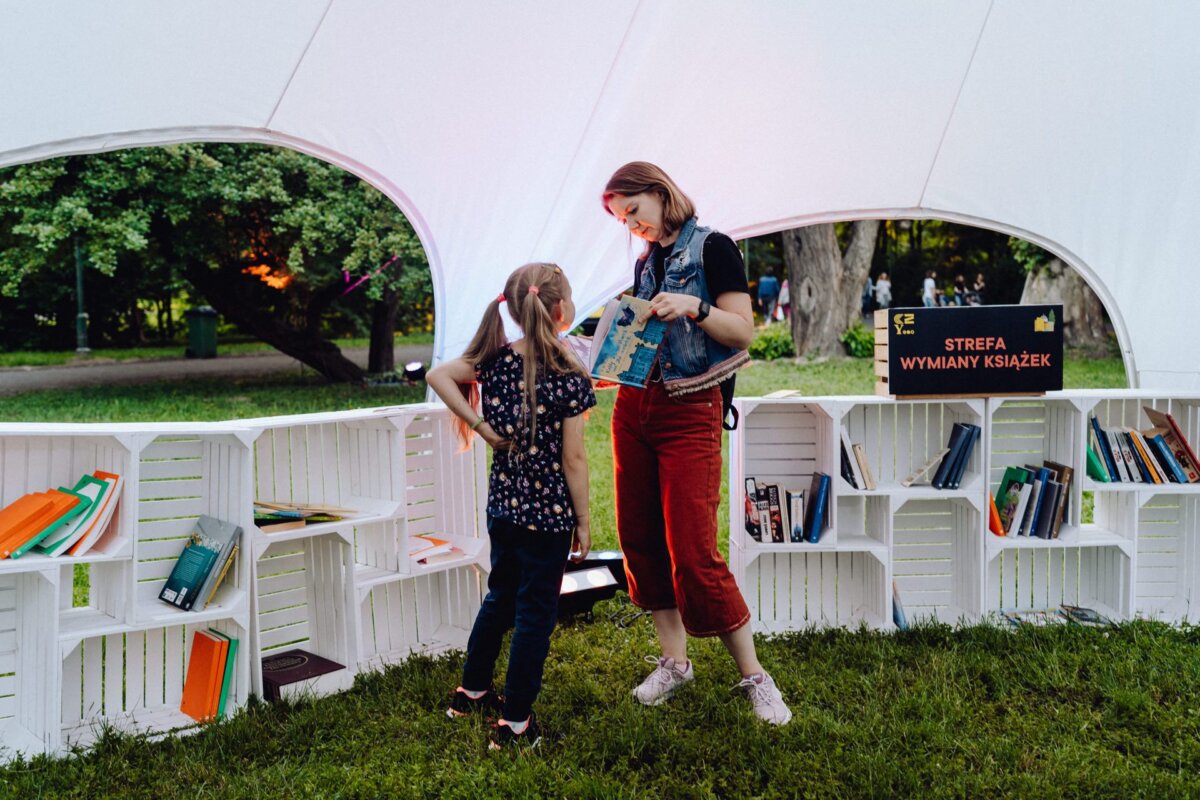 A Warsaw photographer captured a woman showing a book to a young girl in a white tent with open shelves filled with books. The trees are green in the background, and the sign on the tent reads "Book Exchange Zone," or "Book Exchange Zone." This photo report of the event beautifully shows the spirit of knowledge sharing.  