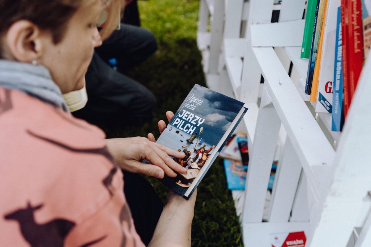 A person in a pink shirt is holding a book by Jerzy Pilch with festive poses on its cover. Standing next to a white fence-like structure with additional books on shelves and green grass in the background, it looks like the perfect event photo captured by an experienced event photographer. 