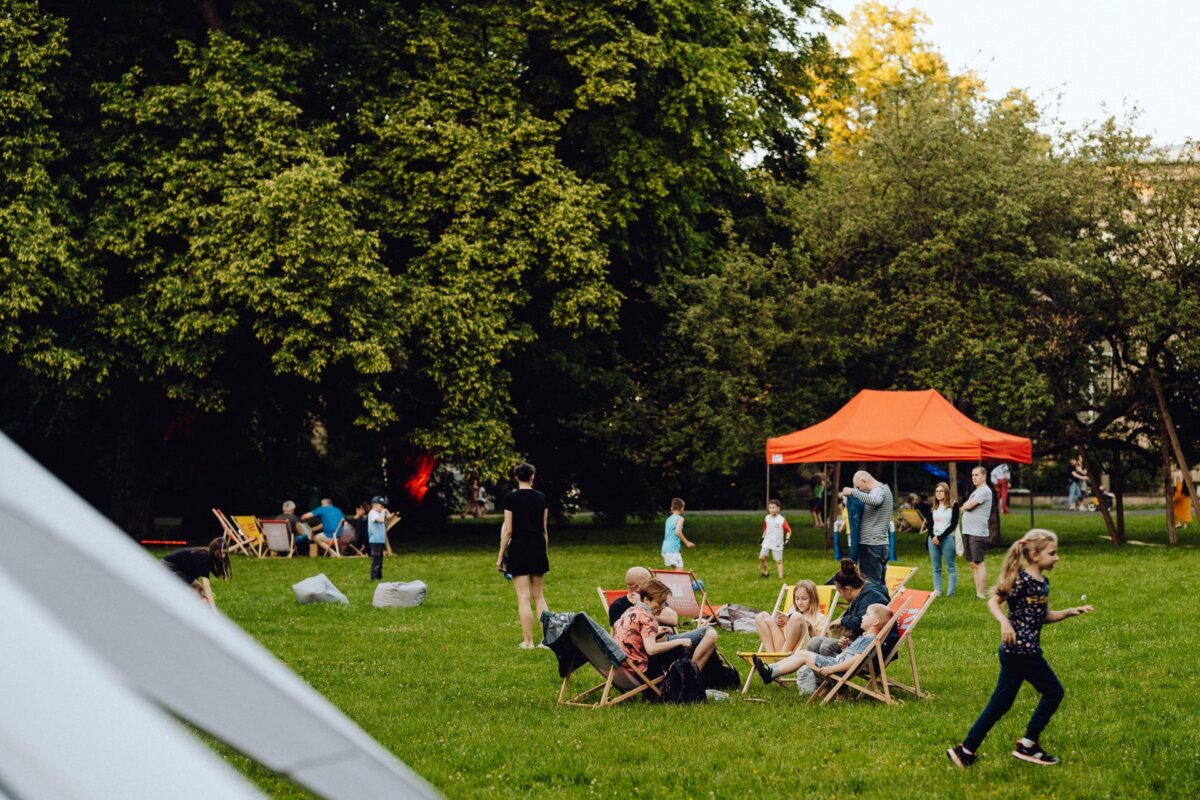 A lively outdoor scene in a park with people relaxing in deck chairs on a grassy lawn. A group of children play nearby, while an orange tent plays in the background. Tall, leafy trees surround the area, creating a natural, inviting atmosphere - perfect for photo coverage of the event.  