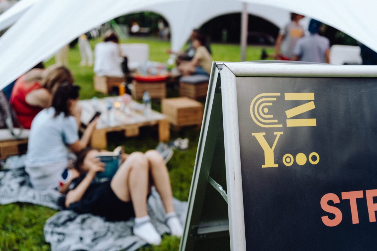 A group of people sit and lie on blankets and wooden pallets under a large white canopy in a park. A signboard with abstract designs and the texts "Yooo" and "STRE" is partially visible in the foreground. The relaxed, casual atmosphere is perfectly captured in this photo report of the event by a photographer from Warsaw.  