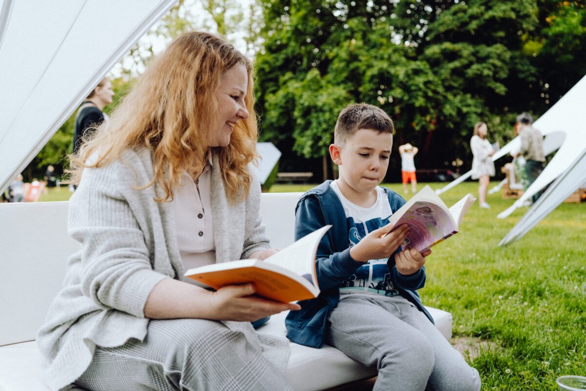 A woman and a young boy are sitting on a white bench outside, each reading a book. The woman has long red hair and wears a bright outfit, while the boy has short hair and wears a blue hoodie. They are in a green park, with a background of trees and other people, beautifully captured by a photographer from Warsaw.  