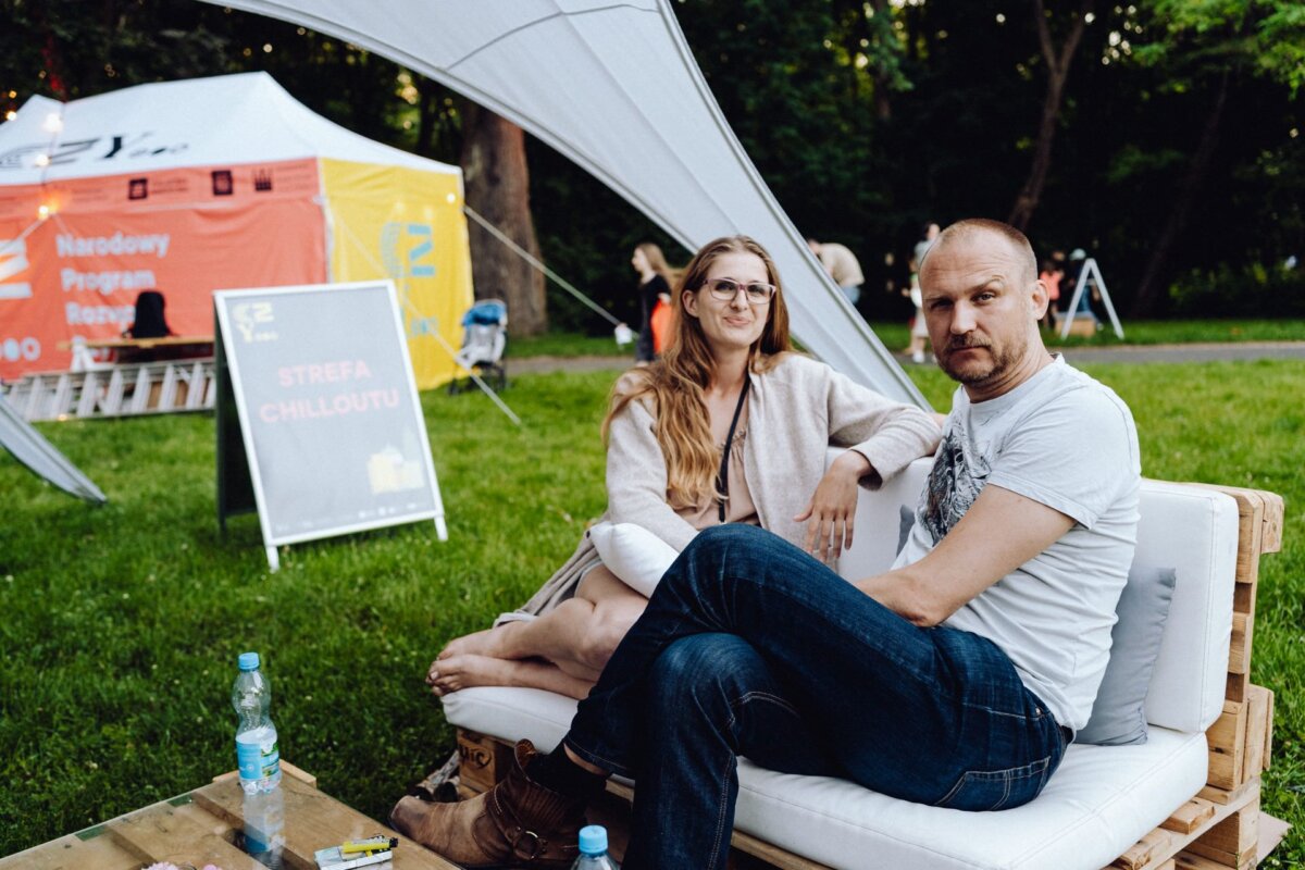 A man and a woman are sitting on a white couch outdoors in a park. The woman with long hair and glasses is dressed in a bright outfit, the man in a white T-shirt and jeans. Behind them is a tent with a sign in Polish suggesting that this may be part of an event captured by the photographer for the event.  