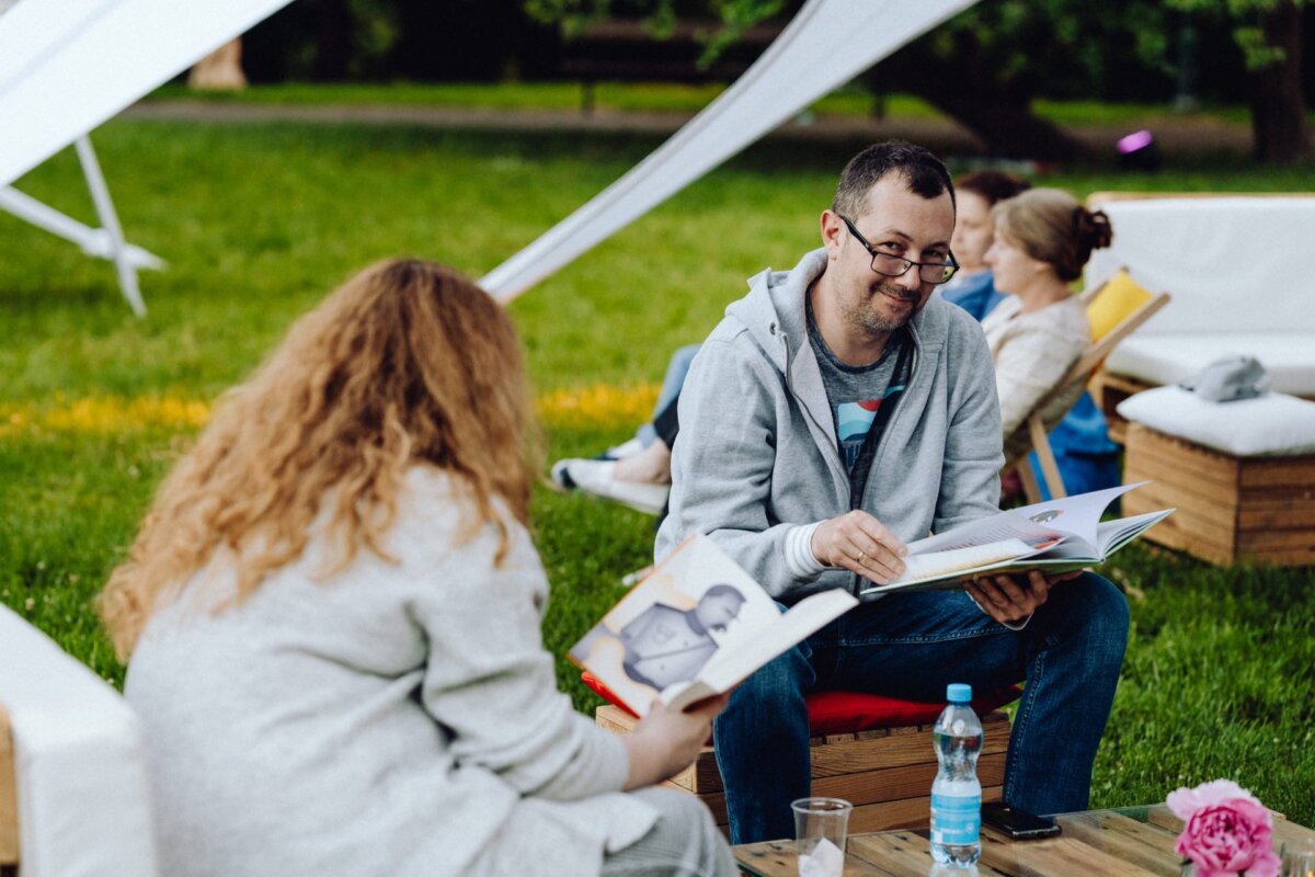 A man and a woman sit outside on wooden chairs and read books. The man with glasses raises his eyes from above his book, while the woman with curly hair focuses on hers. In the background, another person is reading in a chair. A plastic bottle sits on a table, capturing a snippet of life like a photo-op from an event.   