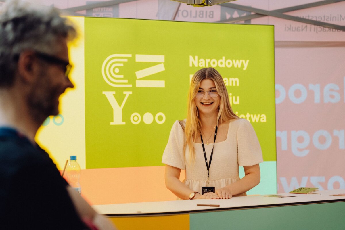 A woman with long blond hair and glasses smiles at a booth with bright, multicolored signage behind her. The text on the signage reads "National" and "Yooo." In the foreground, somewhat blurred, a person wearing glasses, probably an event photographer Warsaw, is visible.  