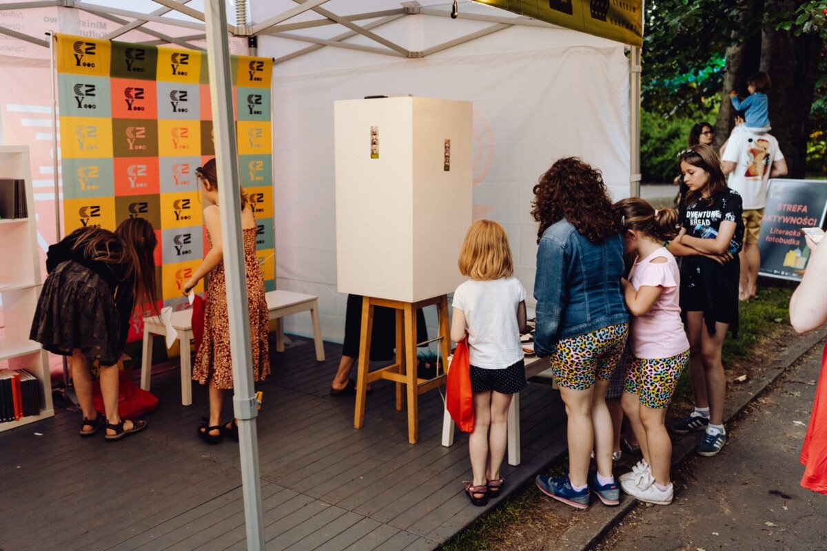 Children and adults gather around an art station in an outdoor tent. Some children pay attention to an invisible activity inside a white box set up on an easel, while others explore their surroundings. In the background you can see a colorful exhibition of posters, perfectly captured by an event photographer warsaw.  