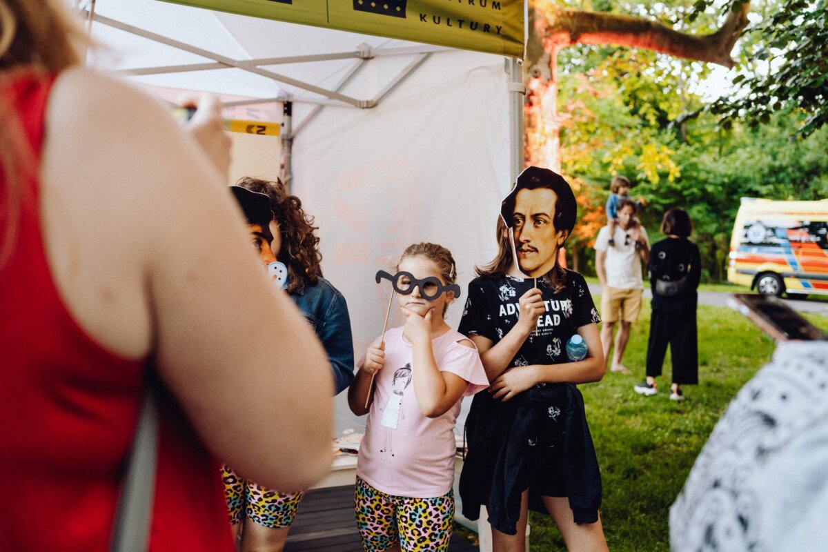 Children standing under a tent at an outdoor event with lush greenery in the background. One child holds a mask resembling a historical figure, while another wears large novelty glasses. The Warsaw-based photographer captured the vibrant scene with other event participants visible in the background.  