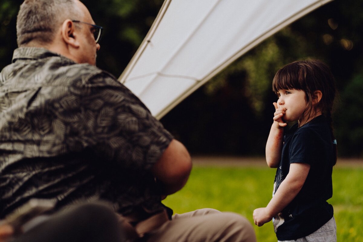 A man in a patterned shirt and sunglasses sits on a chair outside with his back to the camera. A young girl stands in front of him thoughtfully, one hand touching her lips. They are standing near a white tent with green leaves in the background, captured by a photographer from Warsaw.  