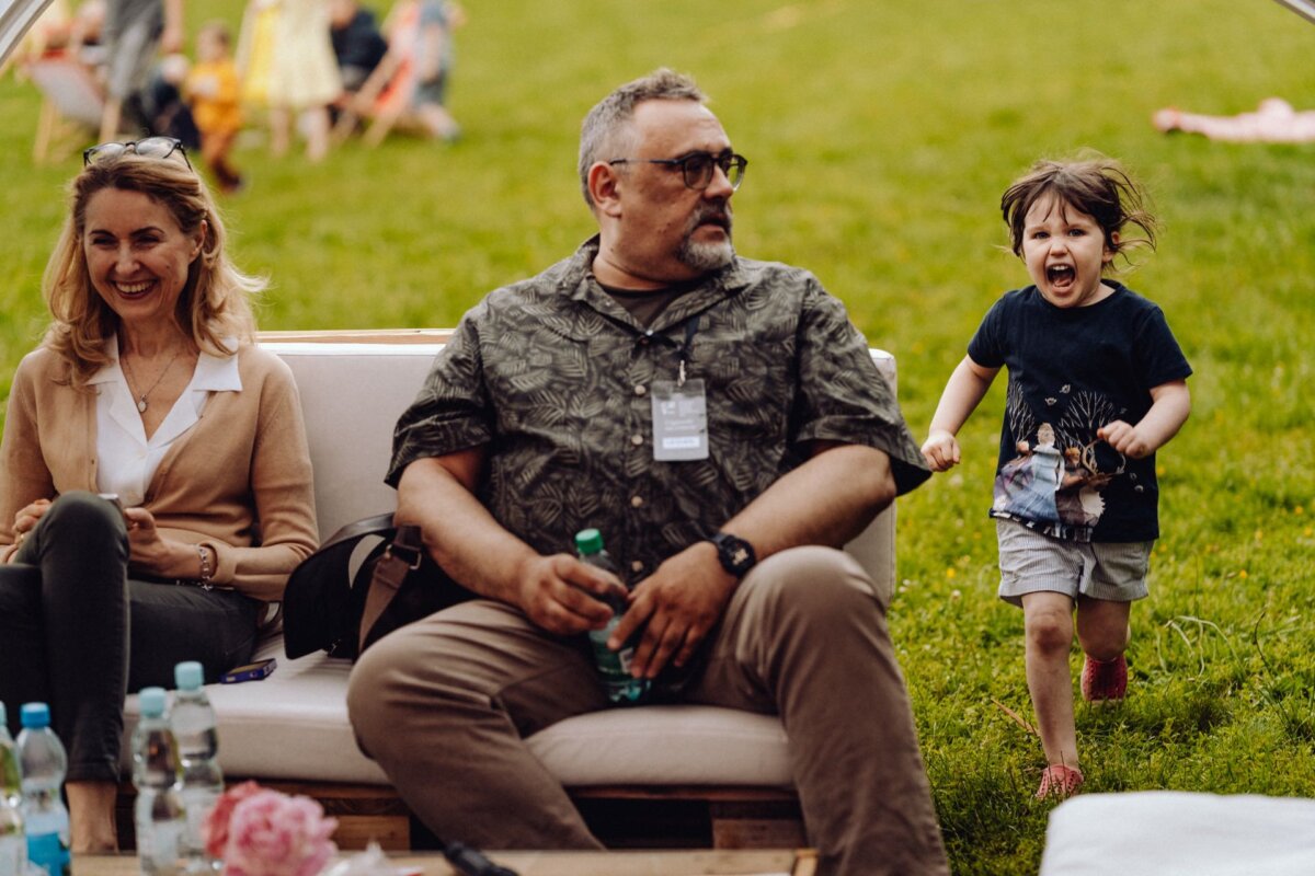 A woman and a man sit on a white couch in the open air, while drinks and flowers stand on a coffee table. A small child runs joyfully beside them on a grassy field. Other people and children can be seen in the background, and the whole thing has been captured by an event photographer warsaw providing authentic photo coverage of the event.  