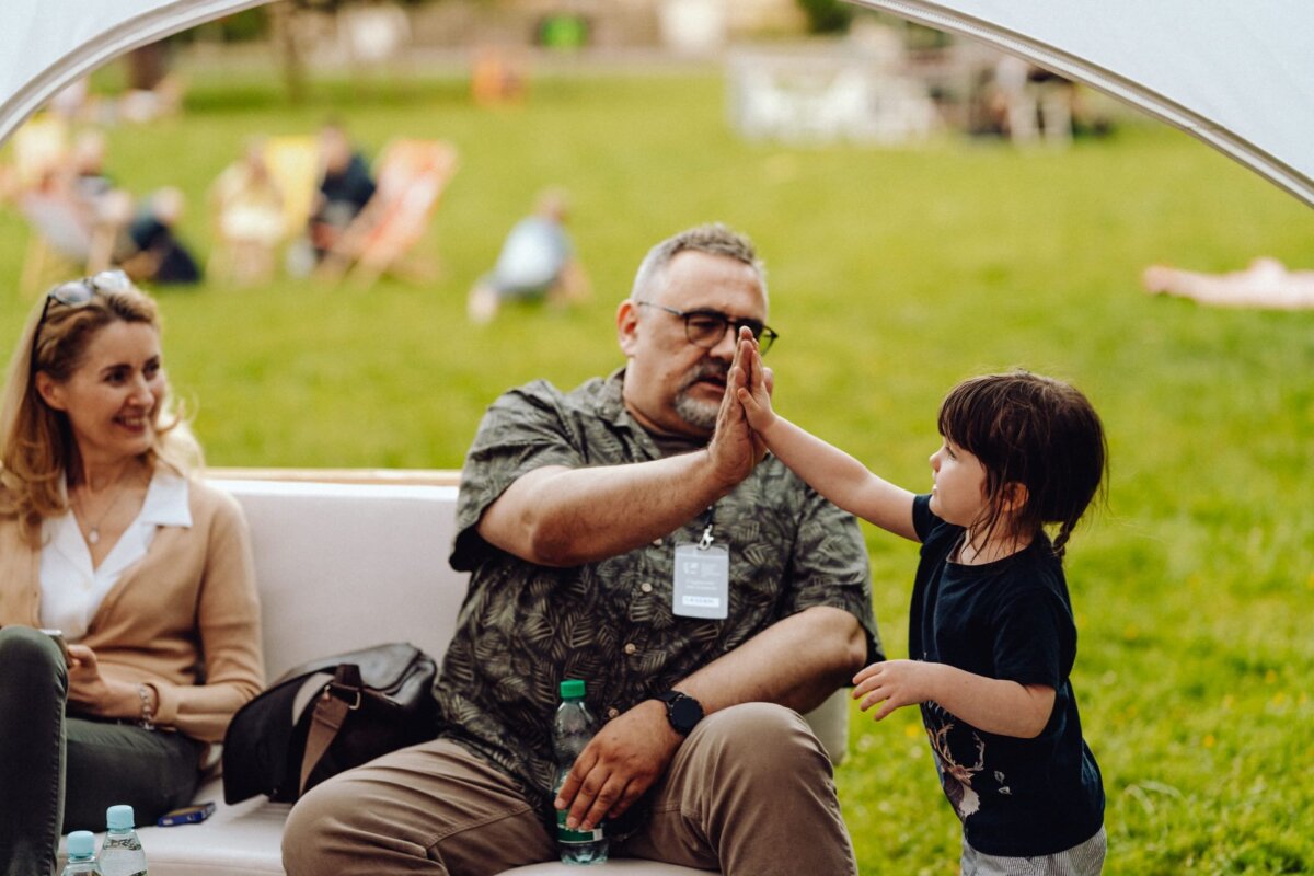 A young child high-fives with a seated man wearing glasses and a name badge in the open air. A woman with blond hair, sitting on the other side, smiles. In the background, you can see people relaxing on the grass - a perfect event photo captured by an event photographer warsaw.  