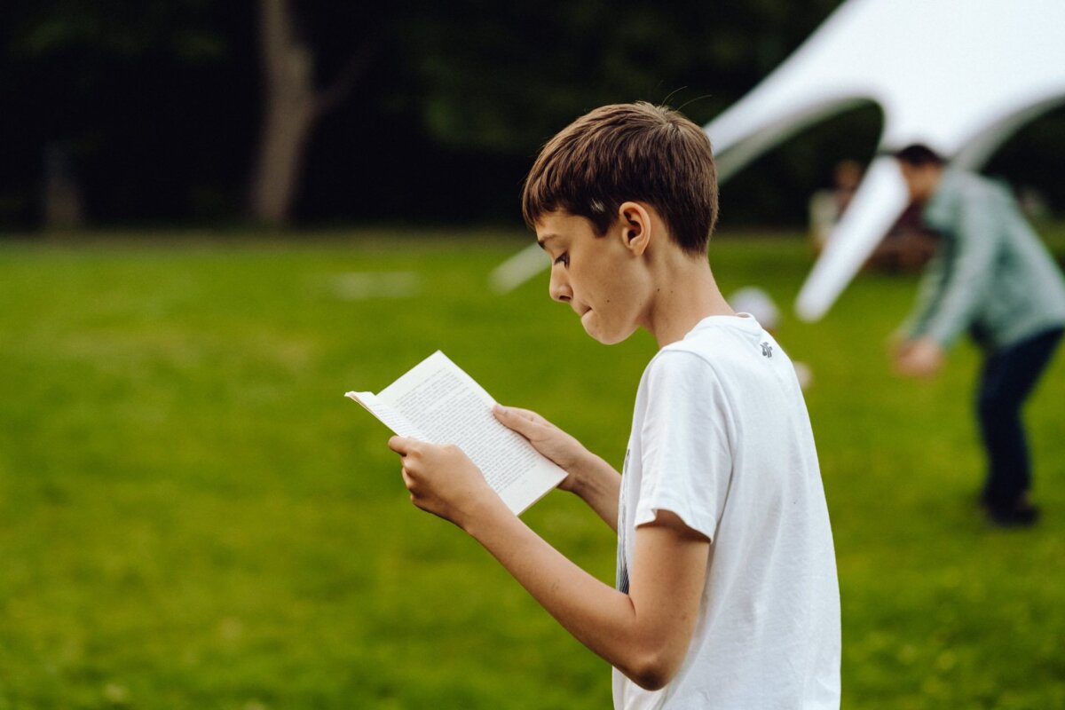 A young boy is standing in a grassy field reading a book, his white T-shirt shining in the sunlight. In the background, captured by the photographer at the event, a person can be seen walking away near a large white tent. The tranquil outdoor area is filled with greenery and trees, adding to this charming event photo.  