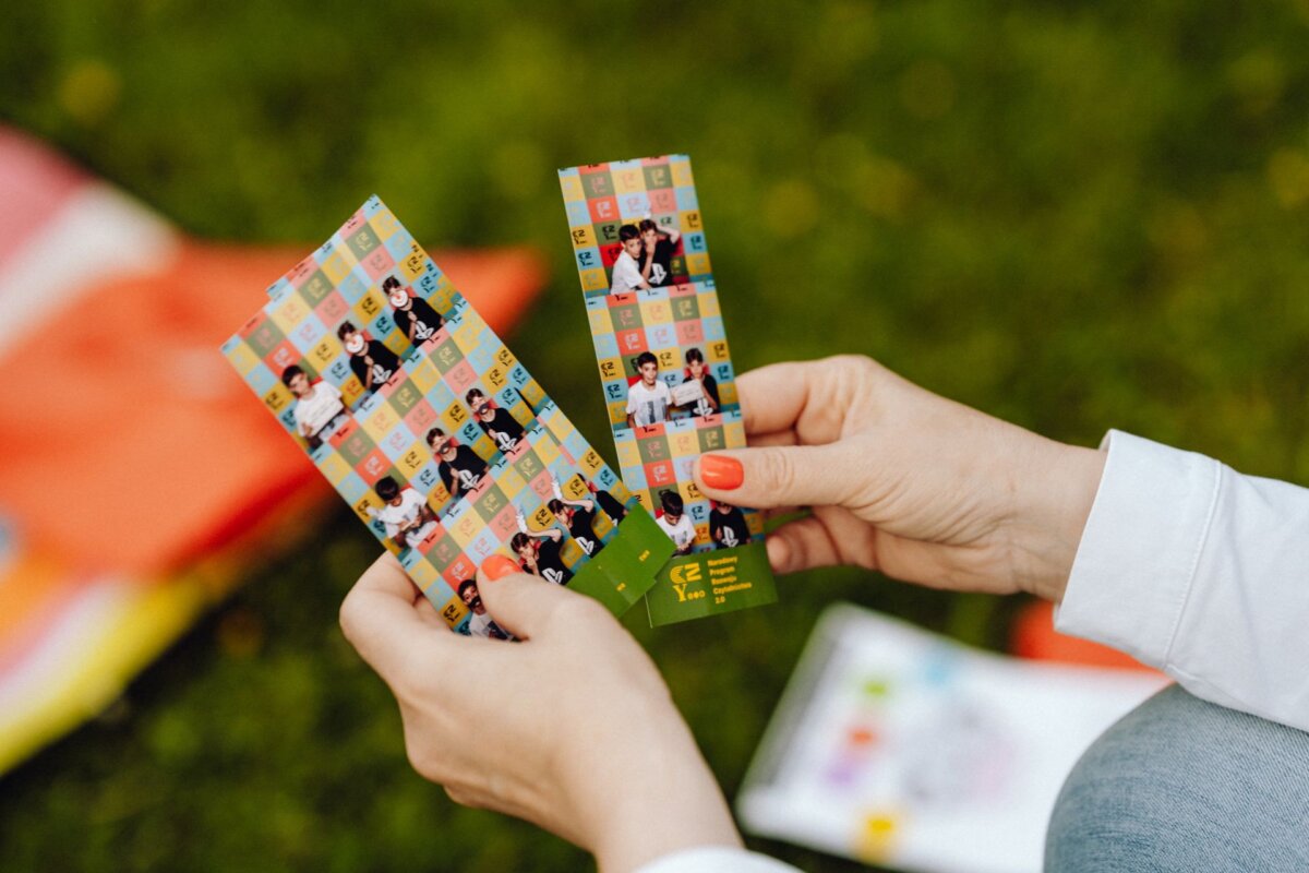 A person with painted nails holds two colorful photo strips with several small photos of smiling people on them. The background is a grassy area, with a fuzzy, open warehouse at the bottom of the image. The person, clearly captured by the photographer from the event in Warsaw, is wearing a white long-sleeved shirt.  