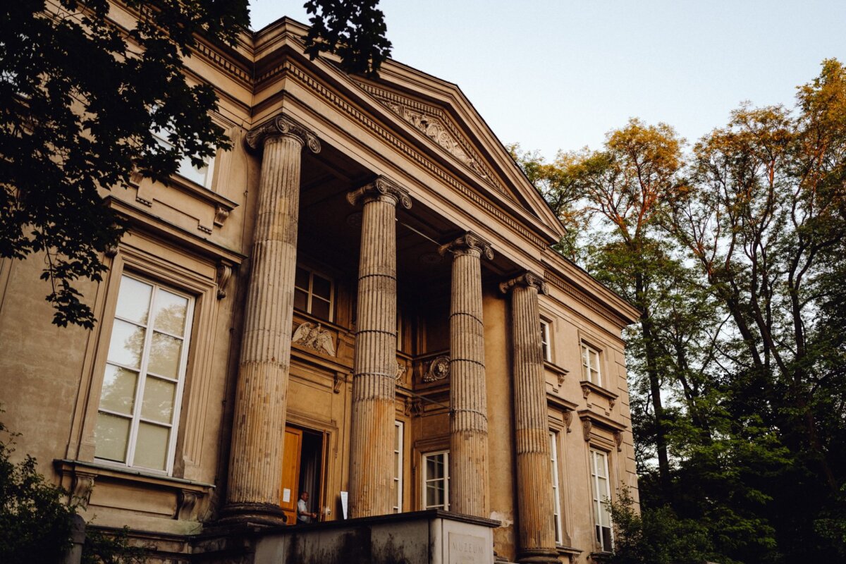 A grand neoclassical building with large columns and ornate architectural details stands majestically. Surrounded by lush green trees whose tops are colored by the setting sun, this picturesque scene was perfectly captured by a photographer from Warsaw. The building is characterized by tall windows and a pediment over the entrance.  