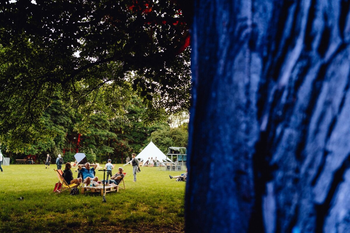 Bright, open field with people sitting at picnic tables under trees. A white tent can be seen in the background, partially obscured by a large blue tree trunk in the foreground. The atmosphere is relaxed, reminiscent of a casual outdoor gathering or festival - perfect for a photography event from Warsaw.  