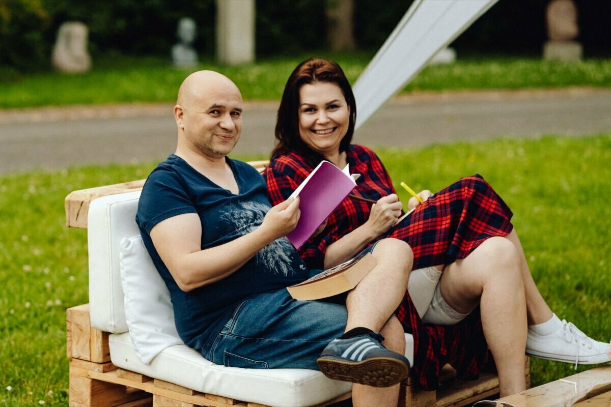 A man and a woman sit on a bench made of wooden pallets in the open air. The man, dressed in a navy blue t-shirt and denim shorts, holds a purple notebook and smiles. A woman in a red checkered dress holds a yellow highlighter and smiles warmly. Grass and trees in the background, captured perfectly for the event photo shoot.   