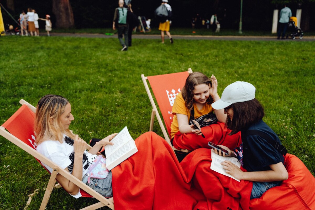 Three people are sitting on red canvas chairs on a grassy lawn. Two are wrapped in blankets, one of them is wearing a white cap. All three are busy talking and holding papers. People are walking in the background, and a baby carriage can be seen - this was captured perfectly by event photographer Warsaw.   