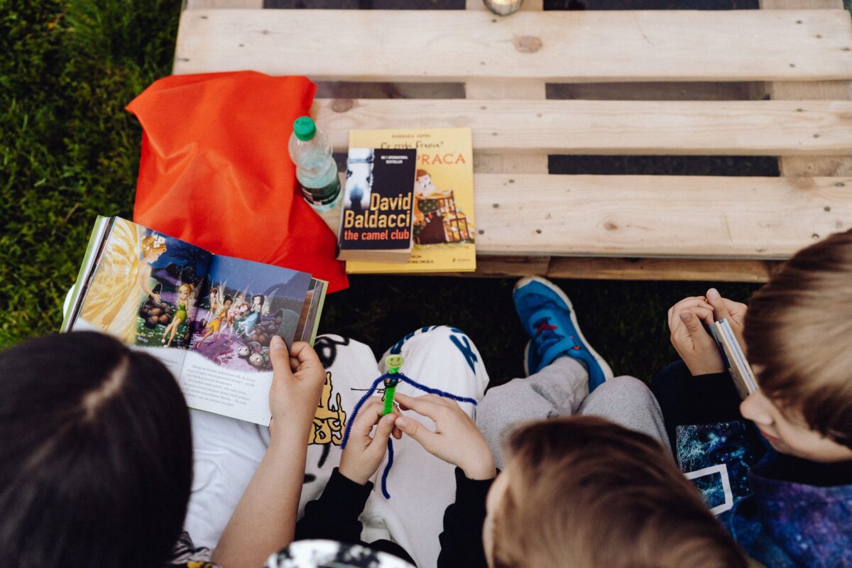 A group of children sitting on a wooden pallet outdoors, reading and looking at books, creates a charming scene perfect for a photo essay of the event. One child is holding a small toy figure. Two books are visible on the pallet: "The Camel Club" by David Baldacci and "Craca." The palette also features a red cloth and a bottle of water.   