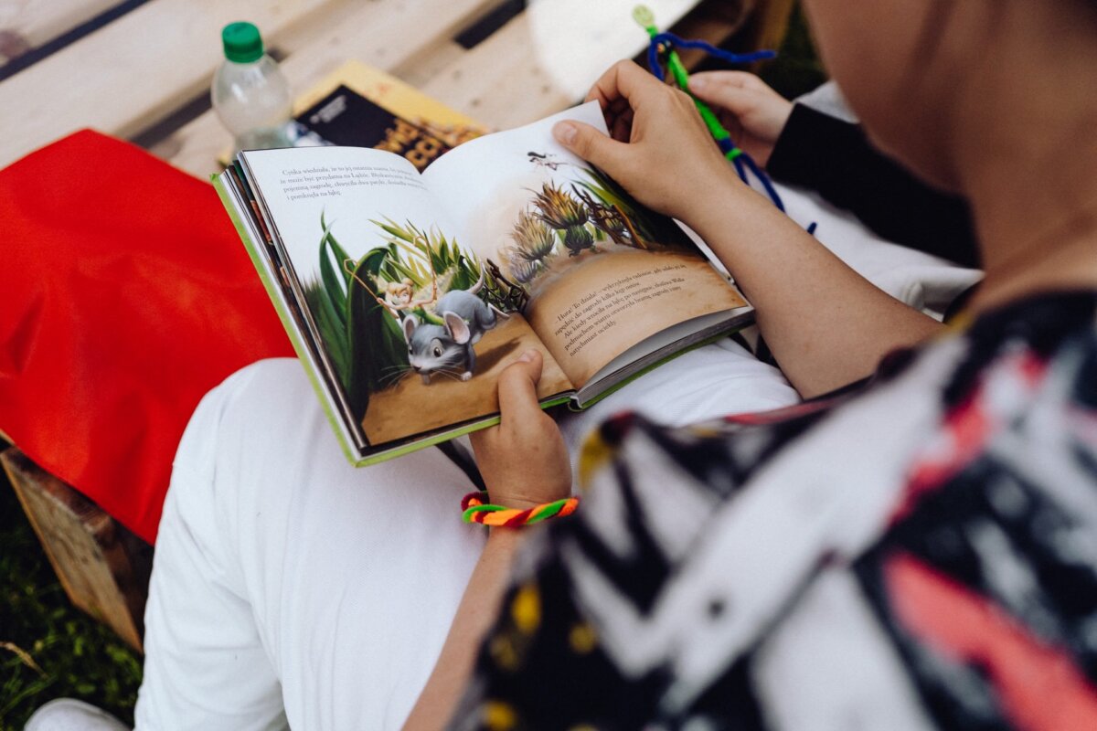 A person reads a children's book with colorful illustrations. The page shows an animated mouse in a garden. The reader, wearing a multicolored bracelet, sits outdoors surrounded by various bags and objects, reminiscent of scenes captured by event photographer Warsaw.  