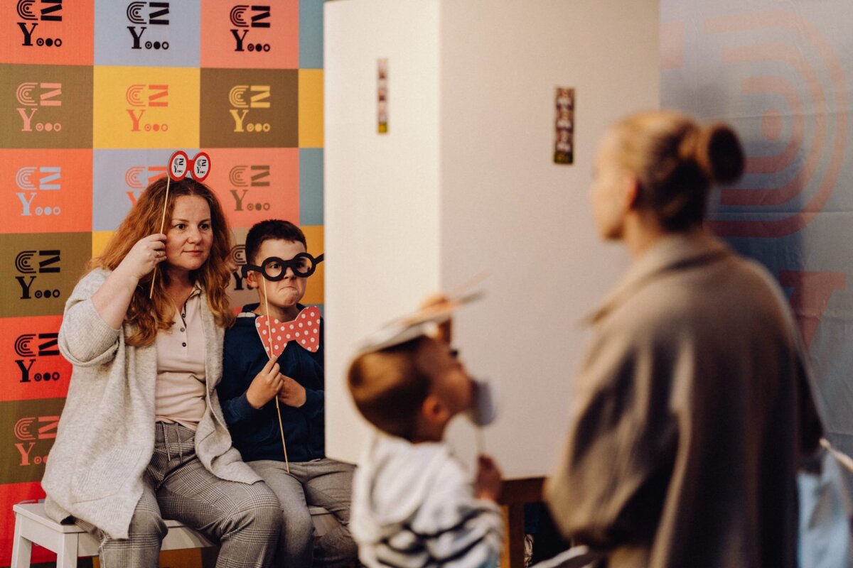 A woman and a boy, both holding funny photo props, smile and pose while a photographer from Warsaw takes their picture. Another child stands in the foreground, holding a mask with a prop on his face. In the background are colorful patterns and the caption "CZYooo".  