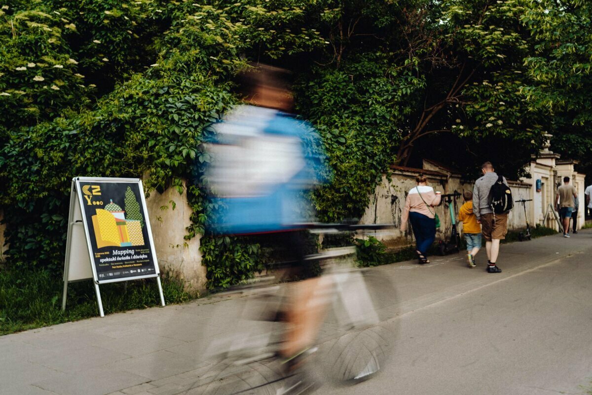 The blurry photo shows a cyclist in a blue jacket riding along a path where people are walking next to a lush green wall. By the wall stands a signboard advertising an event, referring to the work of event photographer Warsaw. Trees and bushes form a dense background.  