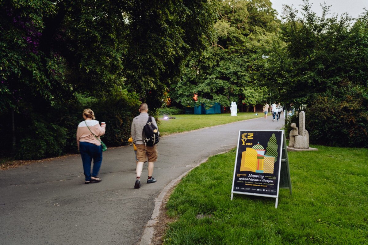 A couple walks along a paved path in a park surrounded by lush green trees. An A-shaped sign next to the path advertises the event with colorful graphics. In the distance you can see other people enjoying the park. If you're looking for an event photographer warsaw, this picturesque area is perfect for capturing memories.   