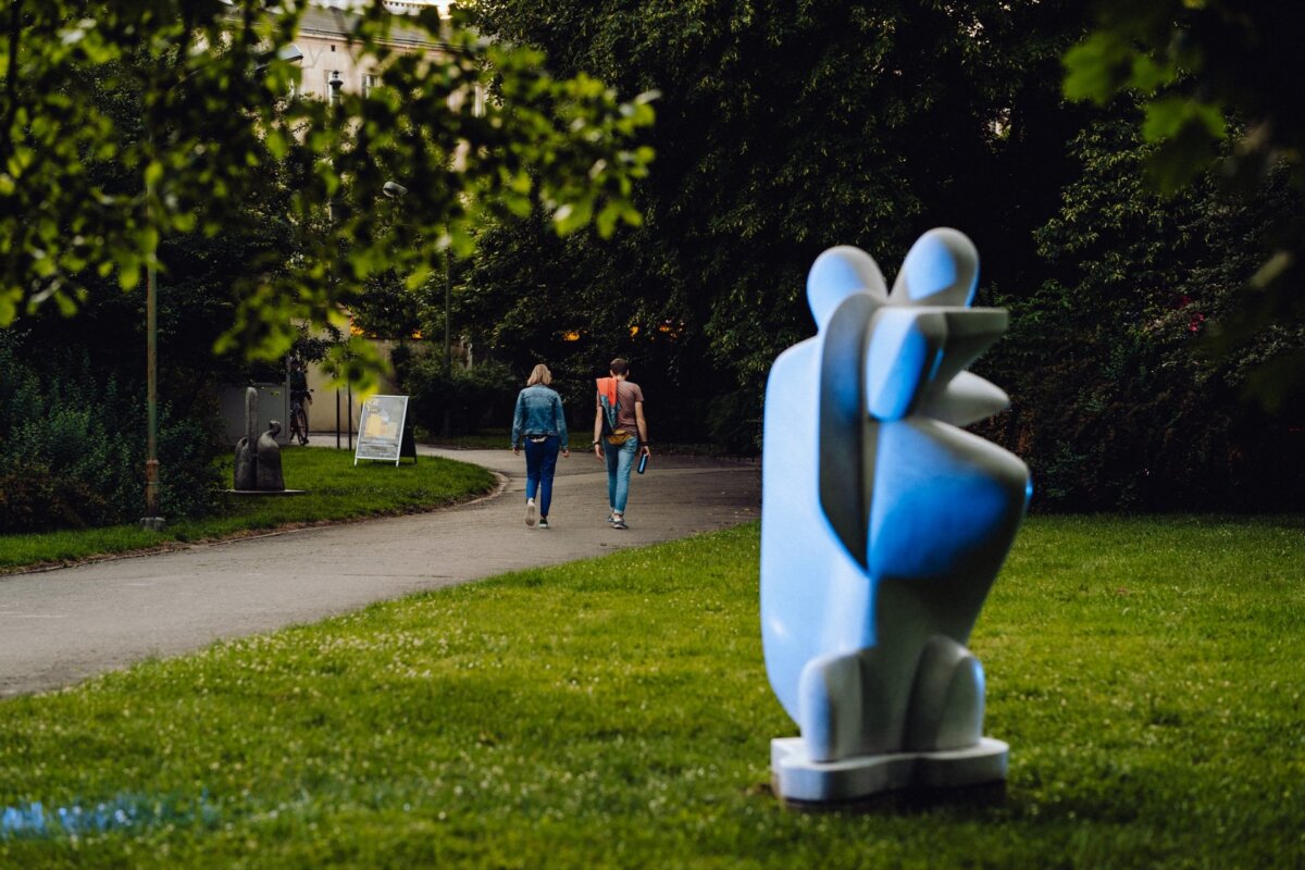 A modern abstract sculpture in pastel blue stands on a lawn in the foreground. Two people are walking along a cobblestone path in the background, surrounded by dense greenery and trees. Along the path, a sign can be seen under the shaded leaves, indicating the perfect moment to take a photo.  