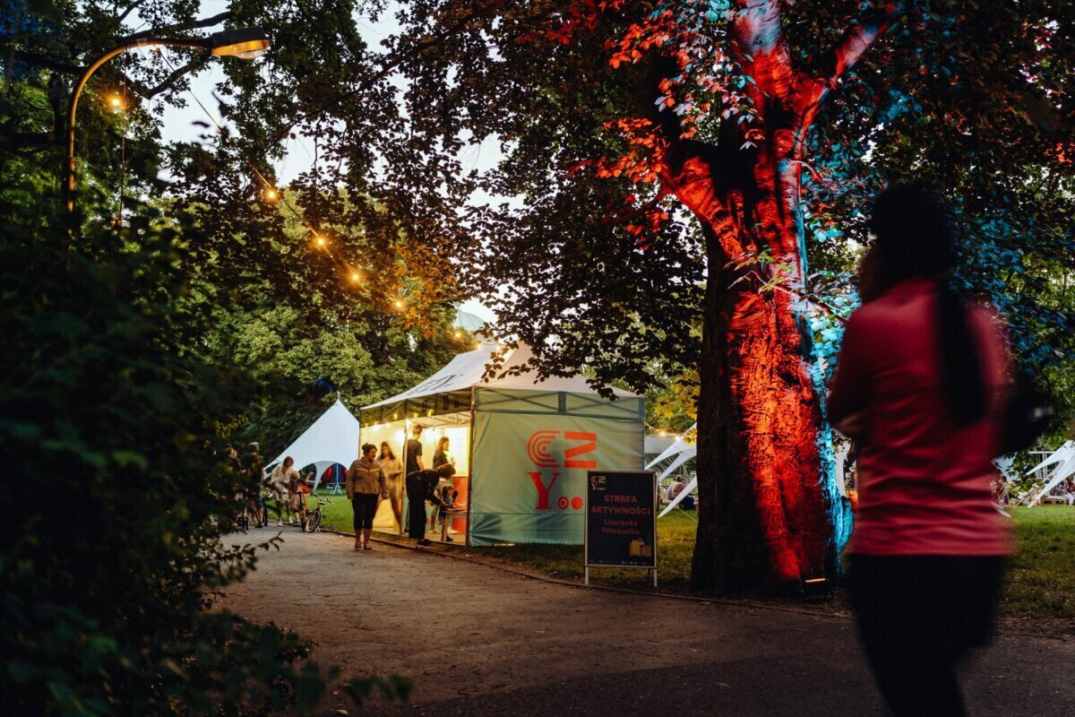 At dusk, a person in a pink shirt walks along a path toward the festival tent. Decorative light chains hang above them, illuminating the area. The multicolored lights highlight the trees, creating a festive atmosphere perfectly captured by the event photographer. In the background, other people are gathering near the tent.   