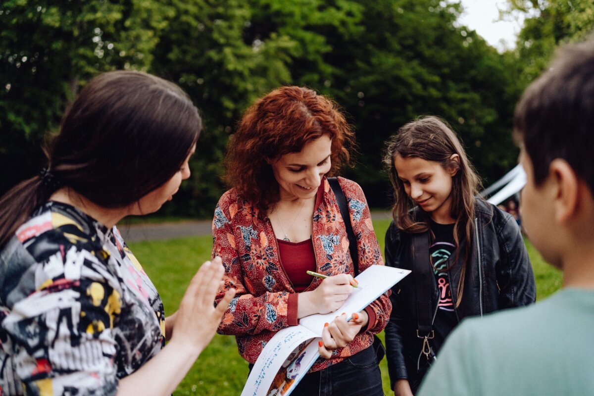 A woman with curly red hair signs an autograph for a young girl, while another girl and a boy stand nearby, smiling and looking on. They are outdoors in a park-like area with green trees in the background, which was beautifully captured by the photographer at the event. 