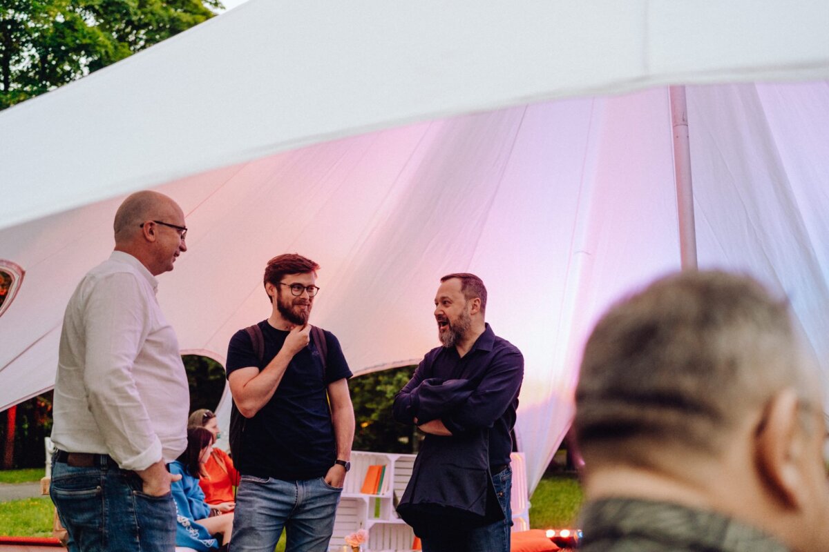 Three men are standing and talking under the large white canopy of a tent at an outdoor event. The man on the left is wearing a white shirt and glasses, the middle man has a beard and glasses, and the man on the right is wearing a dark shirt and has his arms crossed. In this photo report of the event, greenery can be seen in the background.  