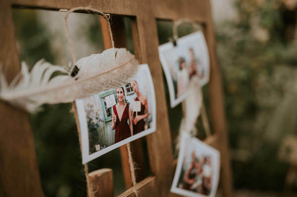 The photos hang on a wooden frame tied with string, decorated with white feathers. The photos captured by Marcin Krokowski show people in various settings. The background is a fuzzy green. The soft, rustic aesthetic suggests a casual party or outdoor celebration.   