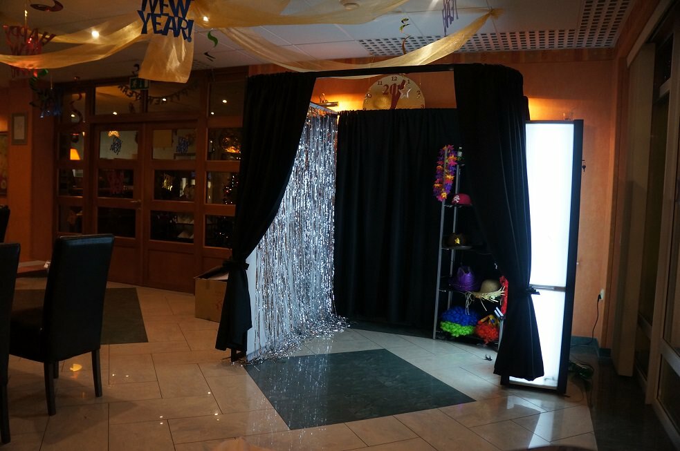 The festively decorated indoor space by event photographer Marcin Krokowski features a photo booth decorated with black curtains and silver candles on one side. A variety of colorful props are visible inside the booth. "Happy New Year" banners hang overhead, suggesting a celebration setting by a well-known photographer in Warsaw.  