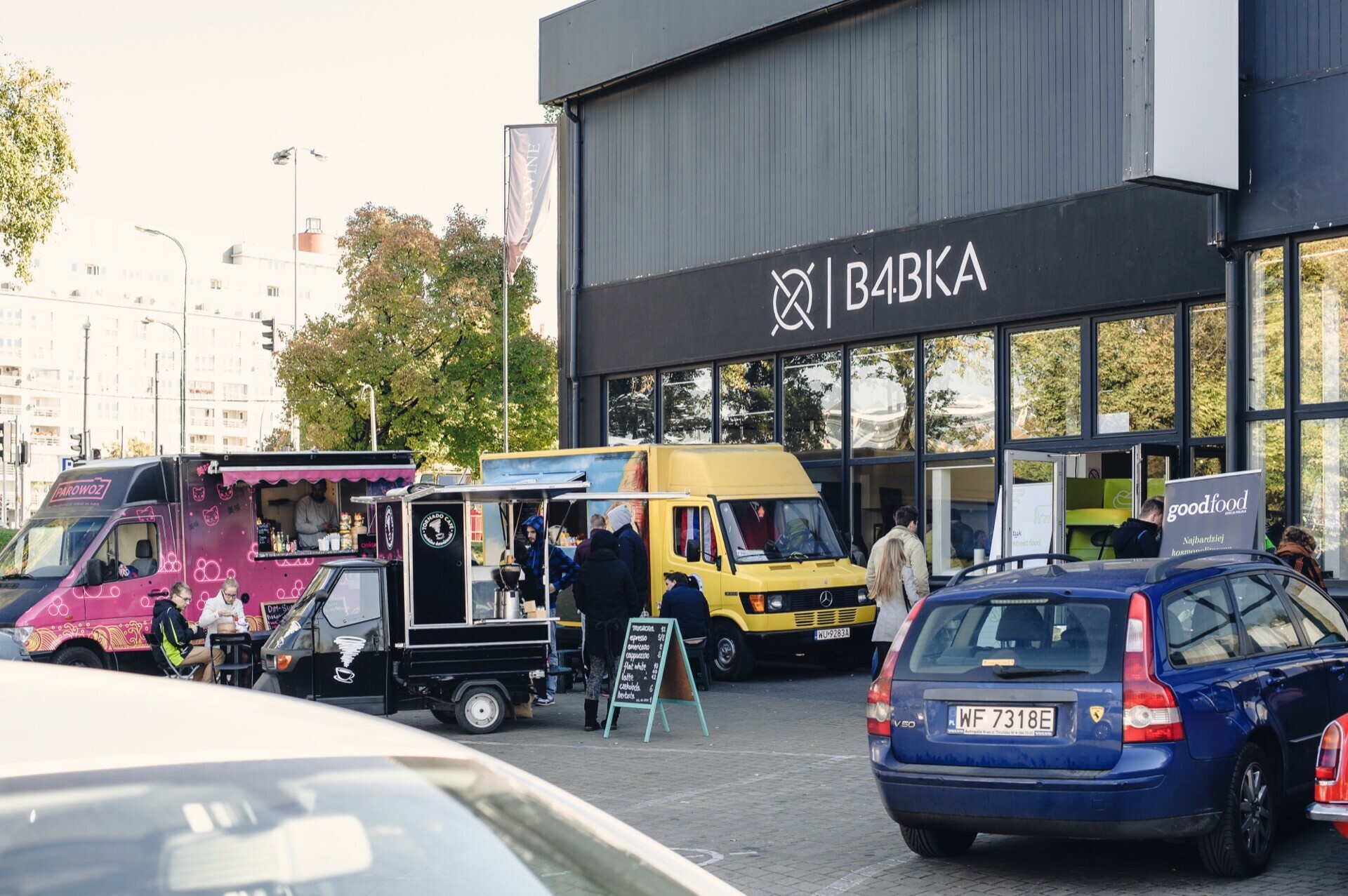 A lively outdoor scene is taking place in front of the building with the sign "Babka." Several food trucks are parked nearby, including a pink one and a yellow one, while people gather for a food fair. A few cars are parked in the foreground, while more people can be seen in the background.  