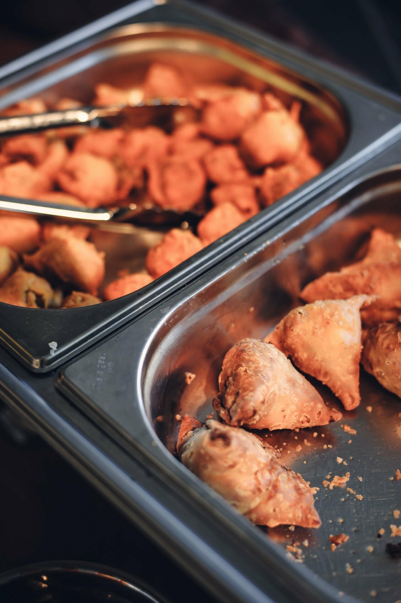 Buffet set with two trays filled with fried snacks, ideal for food fairs. The tray at the back contains small round cakes with metal tongs. The tray in front contains triangular-shaped samosas, slightly golden and crispy.  