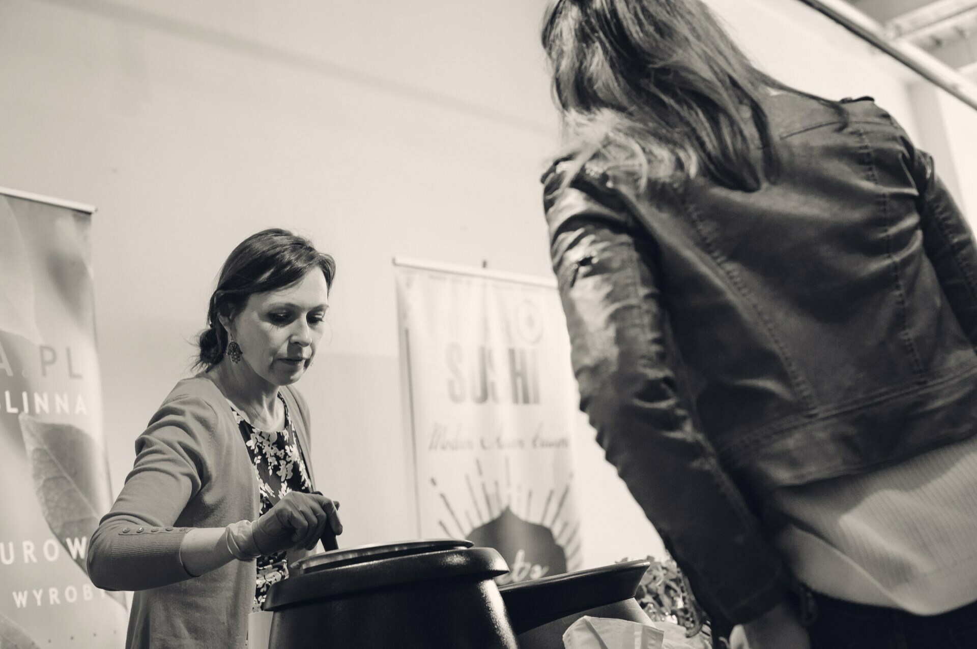 A woman serves food from a large pot at a stall, while another person watches. The scene takes place in a room, as indicated by the surrounding banners and ceiling, suggesting that this may be a food fair. The image is in black and white, which emphasizes the interaction and setting.  