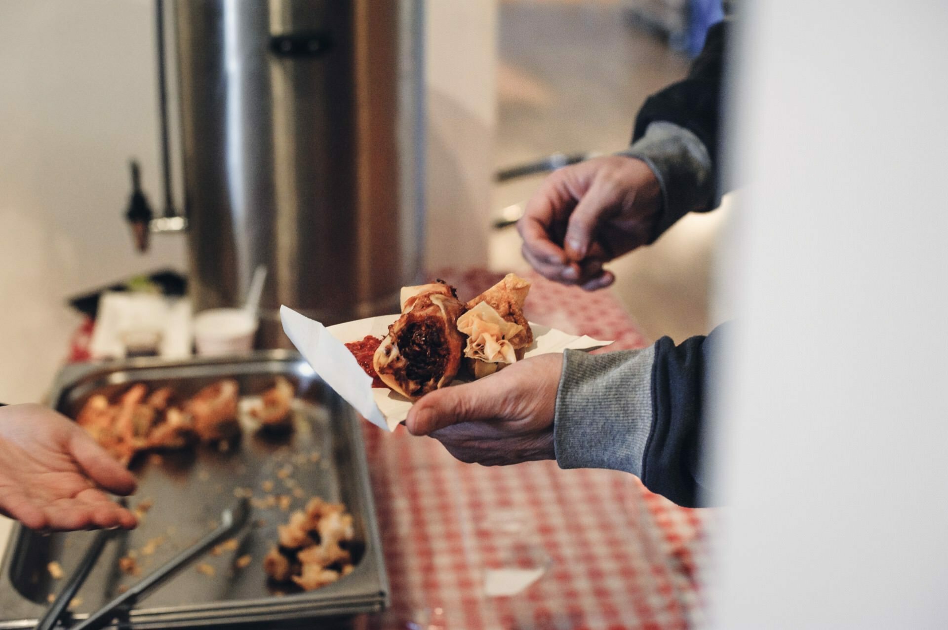 At a food fair, a person holds a paper plate with various baked goods over a checkered tablecloth. Another person, partially visible, reaches out to take a pastry. In the background is a large metal container and a tray with tongs.  