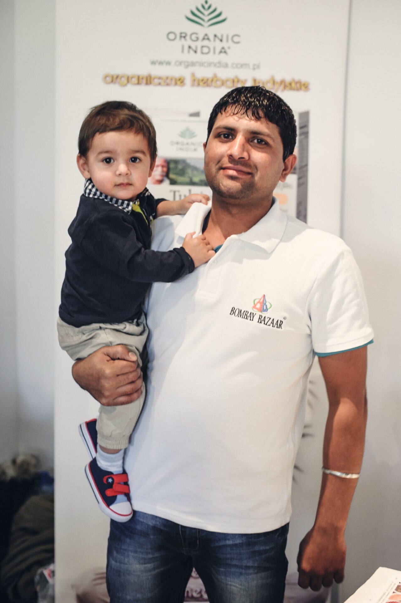 A man in a white polo shirt holds a small child in his arms at a food fair. They are standing in front of a banner with text and logos, including "Organic India." The child is dressed in a dark long-sleeved T-shirt, khaki pants and red and blue tennis shoes.  