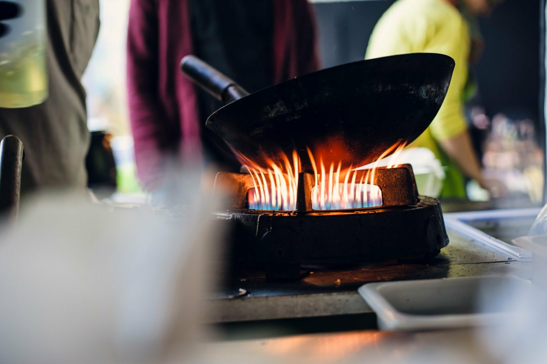 Close-up of a wok on the stove and around it rising flames in the crockery during a cooking fair. Two blurry figures can be seen in the background, one wearing a pink jacket and the other a green shirt, indicating busy cooking. 