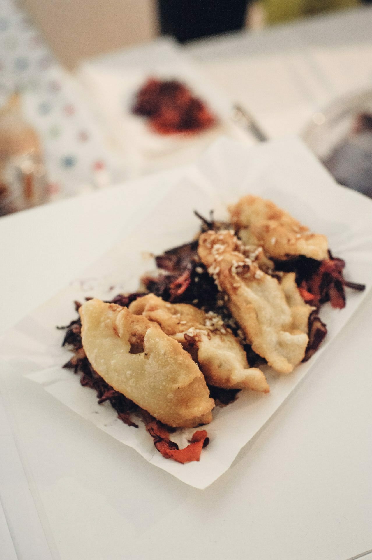 Close-up of a plate of fried dumplings sprinkled with sesame seeds and piled on shredded red cabbage at a food fair. The dumplings appear golden brown and crispy, while other foods and the colorful, patterned background are slightly blurred in the background. 