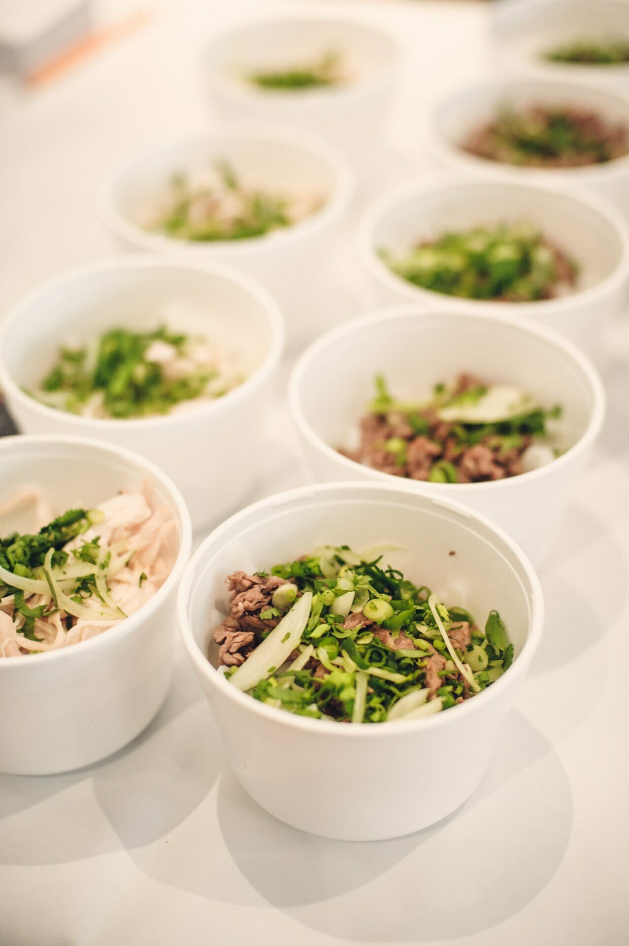 White bowls filled with chopped greens, shredded meat and various herbs are stacked close together on the surface. The ingredients look fresh and give the impression of being part of a prepared dish, such as one intended for a food fair or as a side dish. 