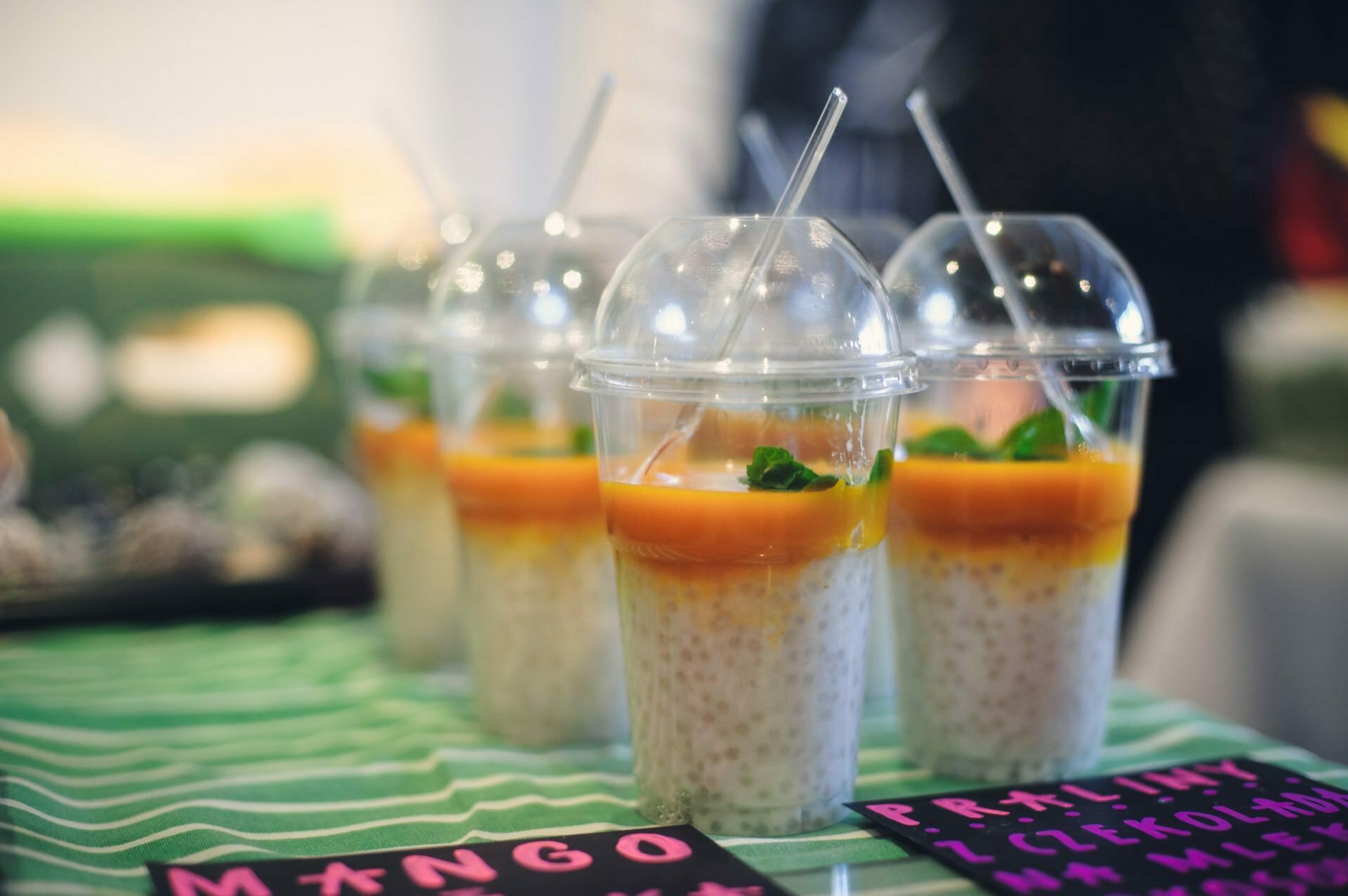 Four clear plastic cups filled with a layer of white tapioca pearls, topped with bright yellow mango puree and decorated with small green herbs. Each cup has a convex lid and straw. Displayed on a table with a green striped tablecloth and handwritten inscriptions, they are perfect for a food fair.  