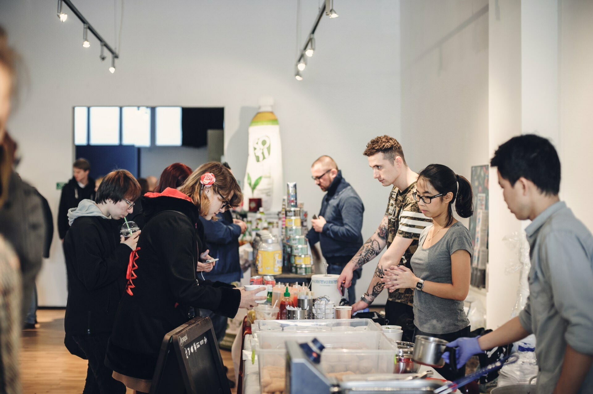 People gather at a modern café or food stand. Some wait in line, others interact with the staff behind the food counter. The interior is bright, with overhead lighting and a variety of food and beverage items on display - the perfect place for a photojournalism event by an author and a photographer for an event.  