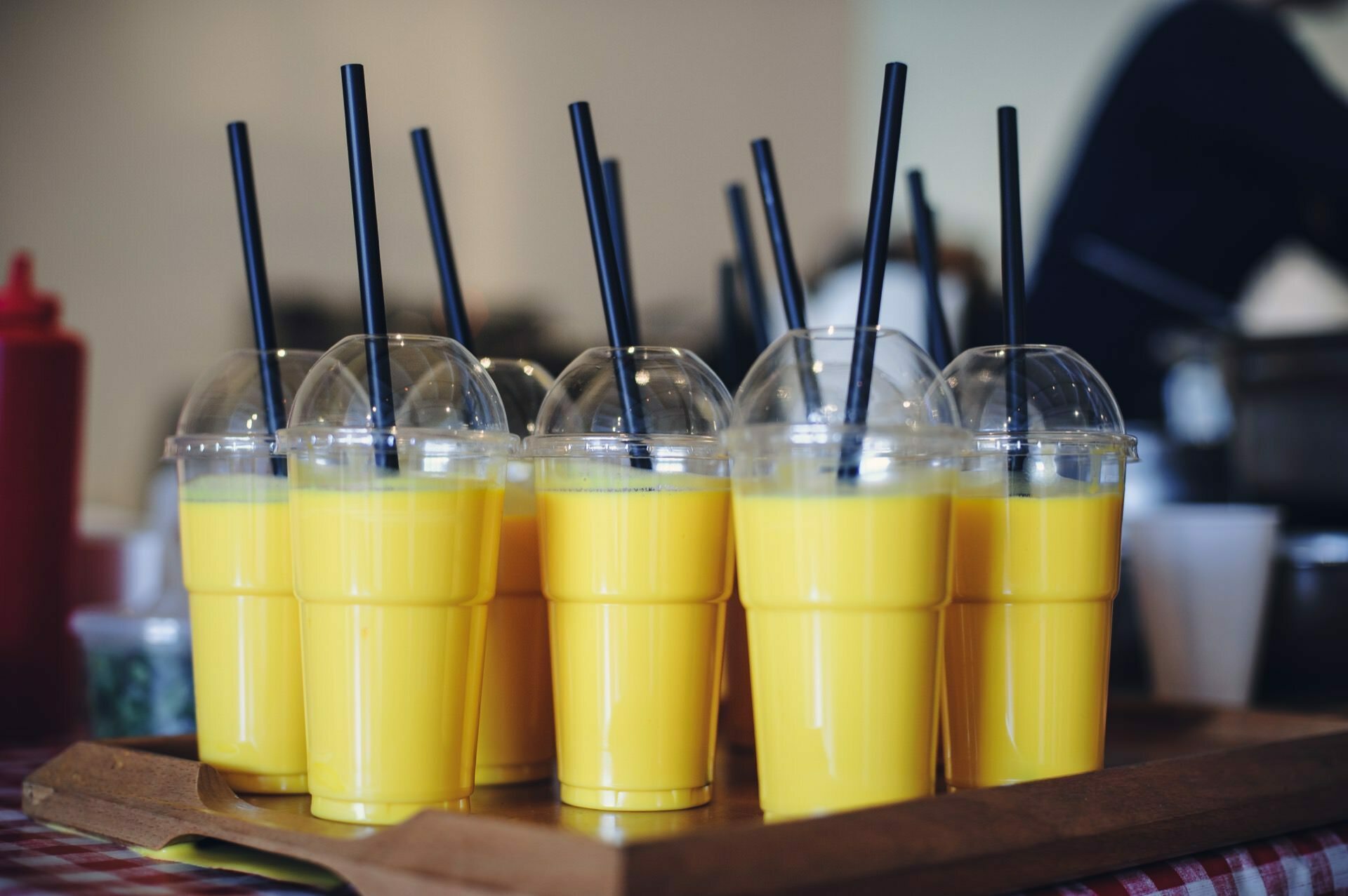 On a wooden tray are several plastic cups filled with bright yellow drinks, each topped with a transparent convex lid and a black straw. The scene suggests a casual setting for a food fair, with a checkered tablecloth and a fuzzy background of condiment bottles. 