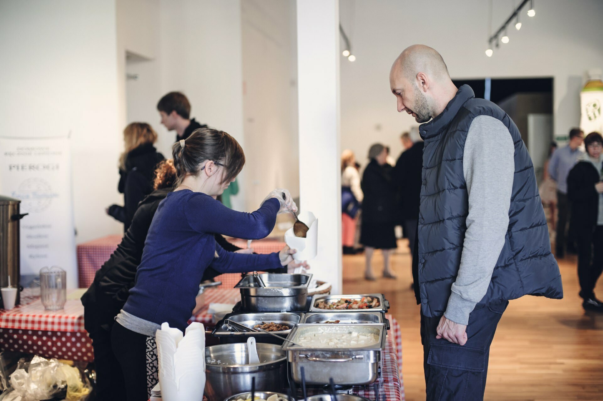 A woman serves food from stainless steel trays at a buffet-style table to a man in a gray vest. The scene, reminiscent of a food fair, takes place in a well-lit room with a wooden floor. People have gathered in the background, immersed in lively conversation.  