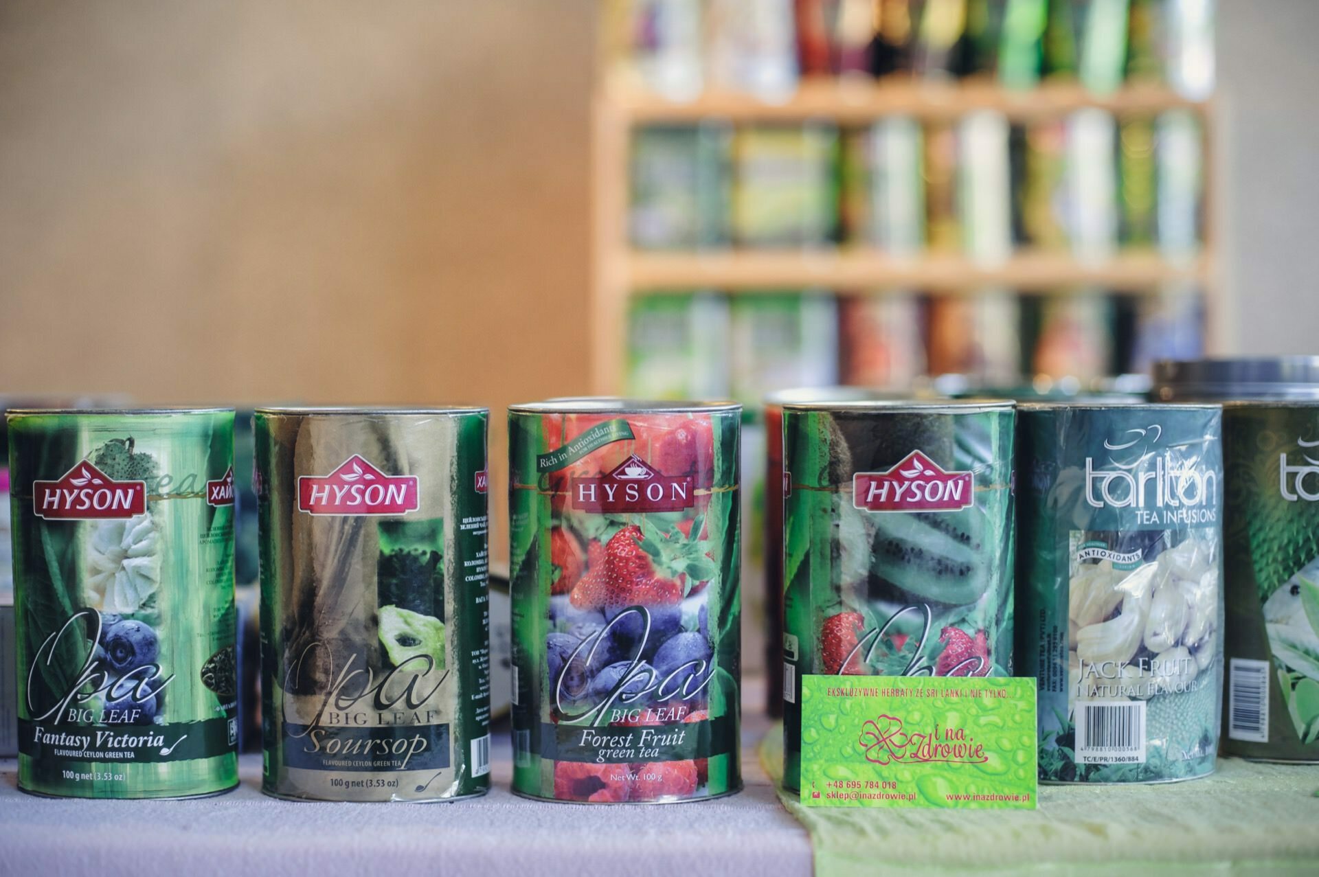 A row of Hyson tea tins is lined up on a table at the culinary fair. The green and black tins contain various flavors, including Fantasy Victoria, Soursop and Forest Fruit. Behind them, a blurry background of more tea products on the shelves can be seen. Herbal tea packaging is also visible.   