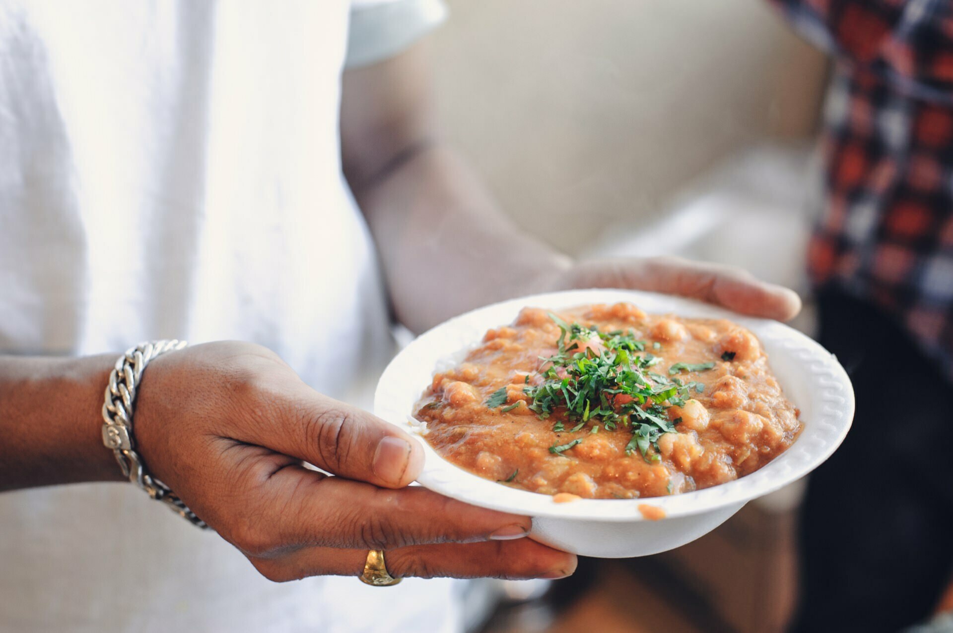 A person wearing a white shirt and silver bracelet holds a white bowl filled with thick stew or soup, garnished with chopped green herbs. The dish looks nutritious, with beans or chunky ingredients visible - this could be a highlight of a food fair. Another person in a checkered shirt is partially visible in the background.  