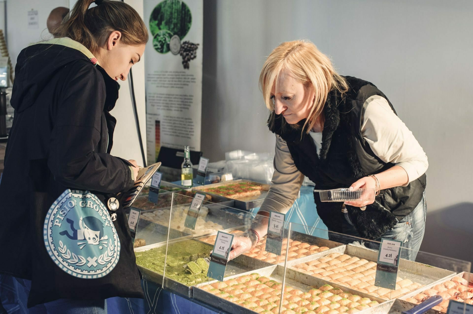 Two women are at a food fair. One with a shoulder bag is watching the selection, holding a notepad. The other, a blonde, selects a sample from trays of neatly arranged green and beige baked goods, possibly cakes or desserts.  