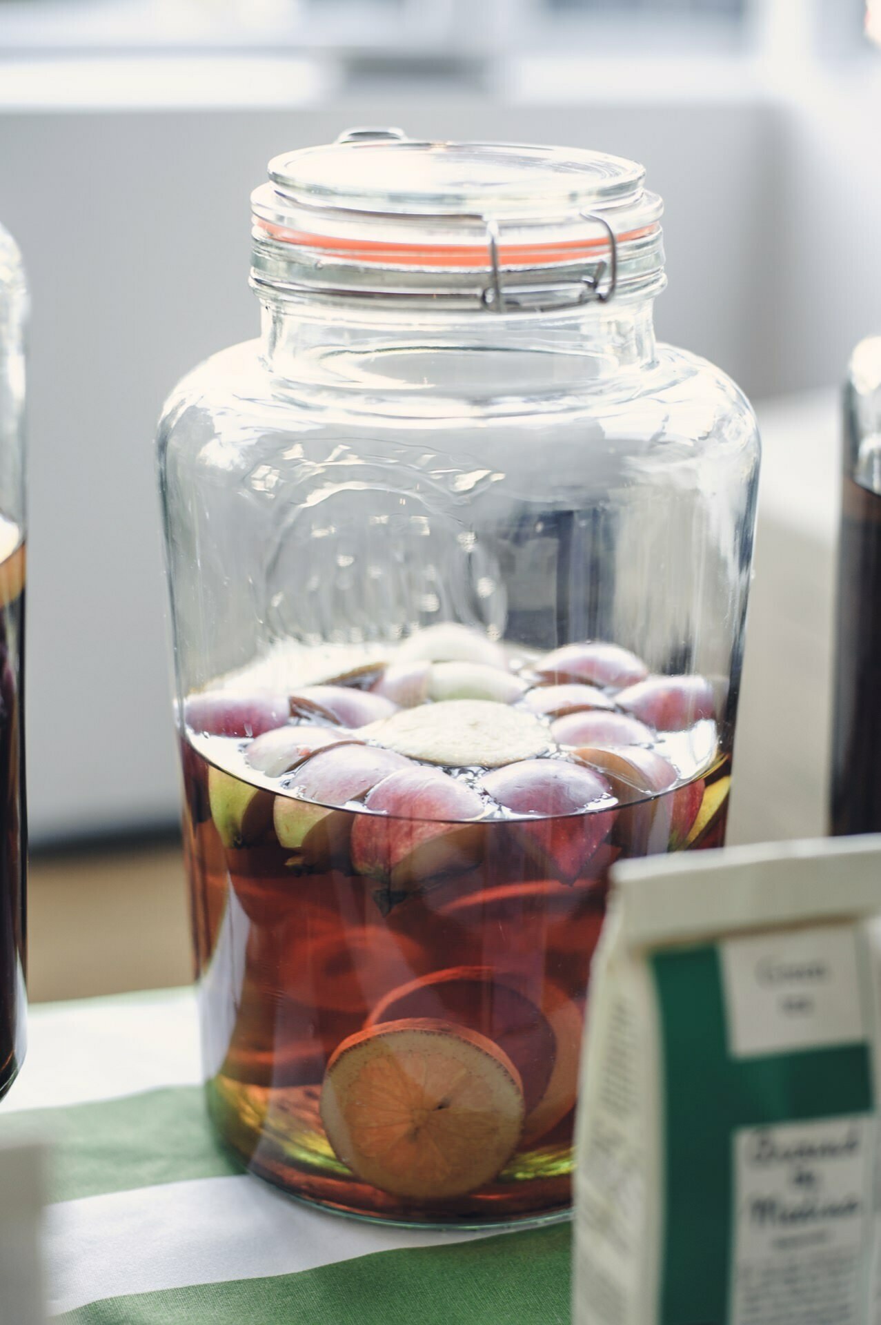 At the food fair, a large glass jar filled with a mixture of liquid and numerous slices of fruit, primarily apples, stands on a table. The jar is closed with a metal latch, and there are other obscure objects nearby, including a box. The scene is brightly lit by daylight.  